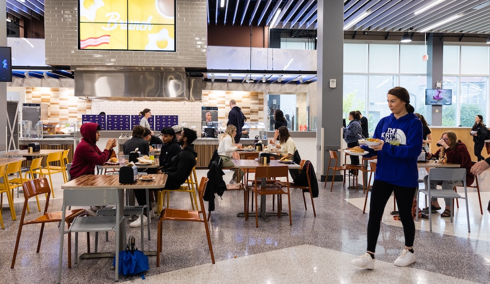 A bustling dining area showcasing various students enjoying their meals at tables, with a food station in the background.