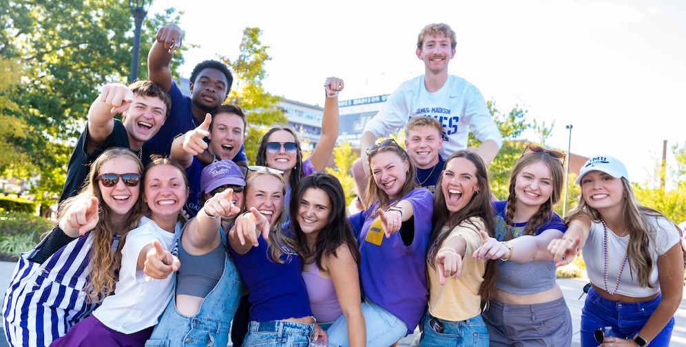 A group of enthusiastic college students posing together outdoors, all pointing and smiling, wearing purple and white attire.