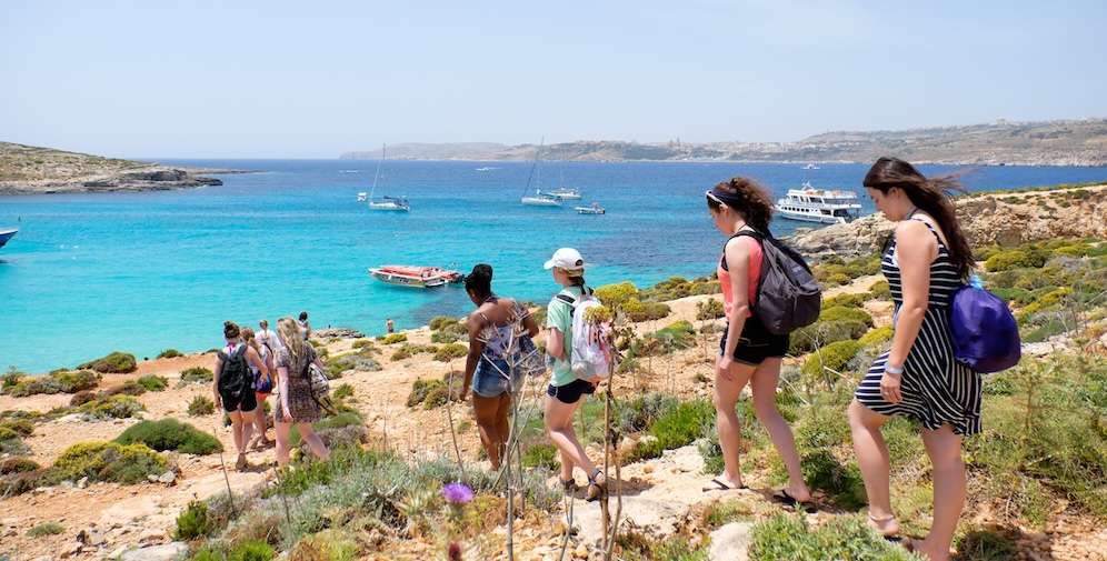 A group of friends hiking down a rocky path towards a bright turquoise sea with boats in the background.