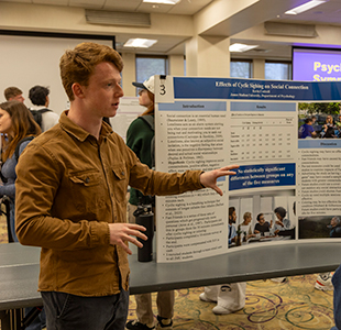 A student discusses their poster about cyclical sighing and social connections with a symposium attendee.