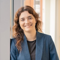 Maggie Testerman smiling with wavy brown hair wearing a blue blazer, leaning against a wall in a modern indoor setting.