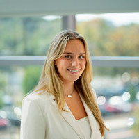 A smiling young woman in a purple polo shirt stands outdoors with greenery in the background.