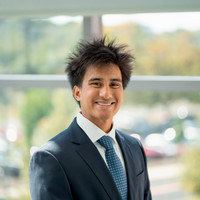 A smiling young man in a purple polo shirt stands on a balcony, surrounded by greenery.