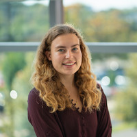 A smiling young woman with curly hair wearing a purple shirt stands outdoors against a backdrop of greenery.