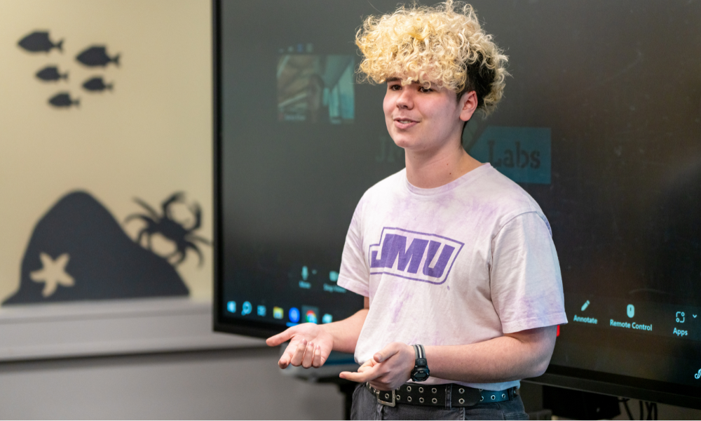 A student with curly hair is wearing a JMU t-shirt and presenting in front of a TV screen displaying the X-Labs logo.