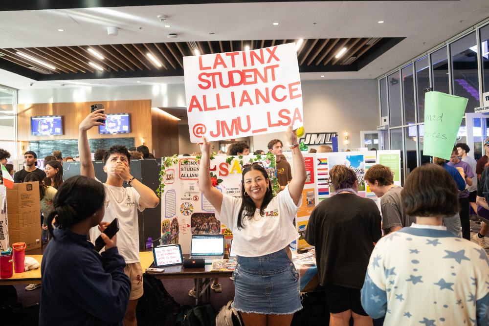 a student holds up a sign reading "latinx student alliance @jmu_lsa" in front of a table for the group. Other student org tables are in the background