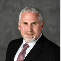 Image of a smiling white man with greying hair and blonde & grey goatee, in a black suit, white shirt and redish tie, in front of a dark grey background.