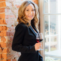 Headshot of Noelle Allen, a sideview of a smiling blonde woman in a dark suit holding a glass of red wine, standing in front of a brick wall and window