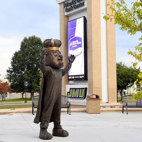 A bronze statue of duke dog wearing a crown, standing with arms raised, located in front of the Atlantic Union Bank Center at JMU.