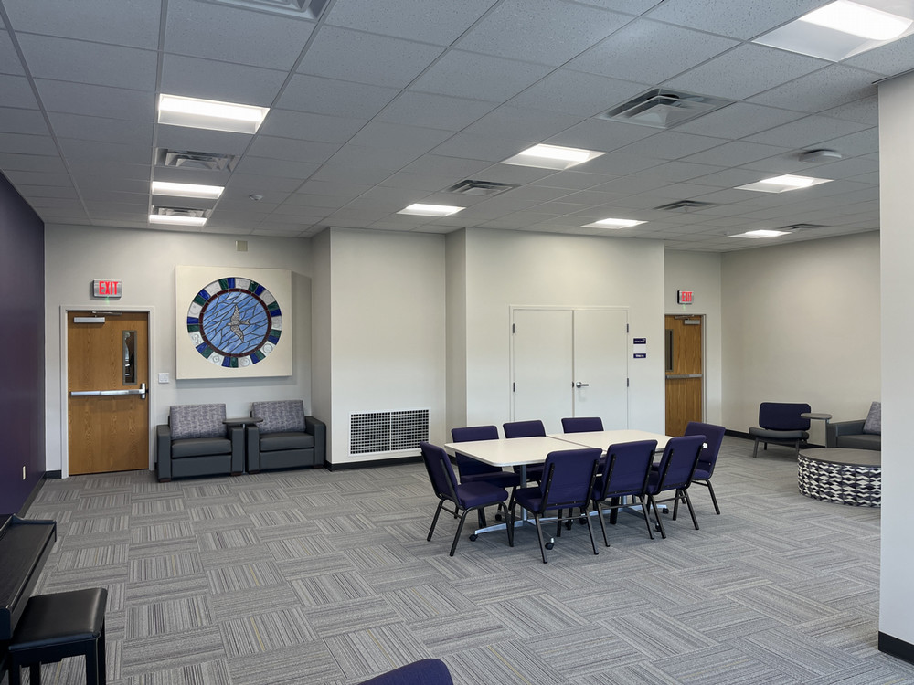 a view of the interfaith space centered on a rectangular table with 8 chairs around it facing the front wall and the room exits