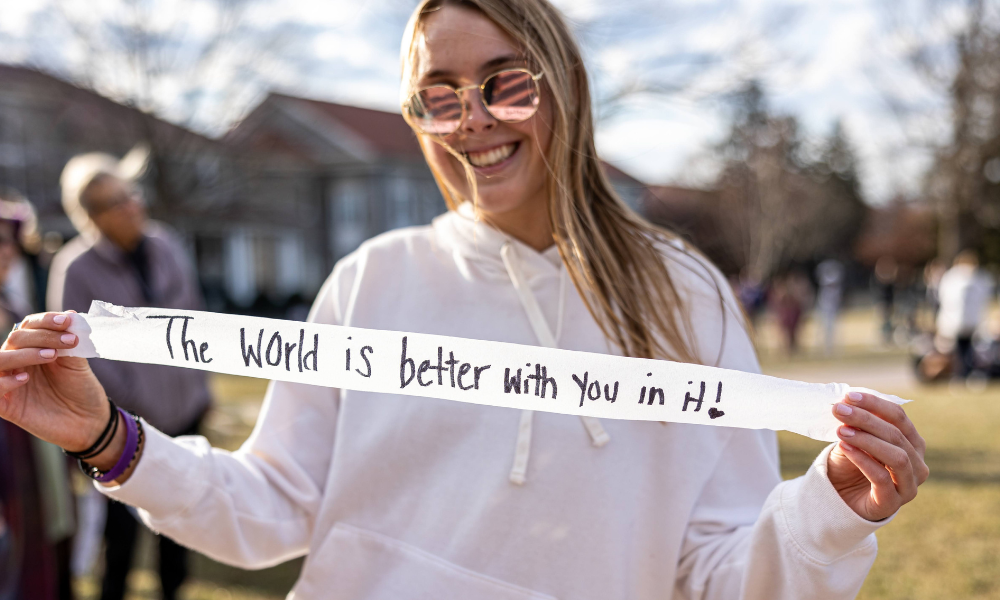 Student holding a message on a streamer that reads: The world is better with you in it!