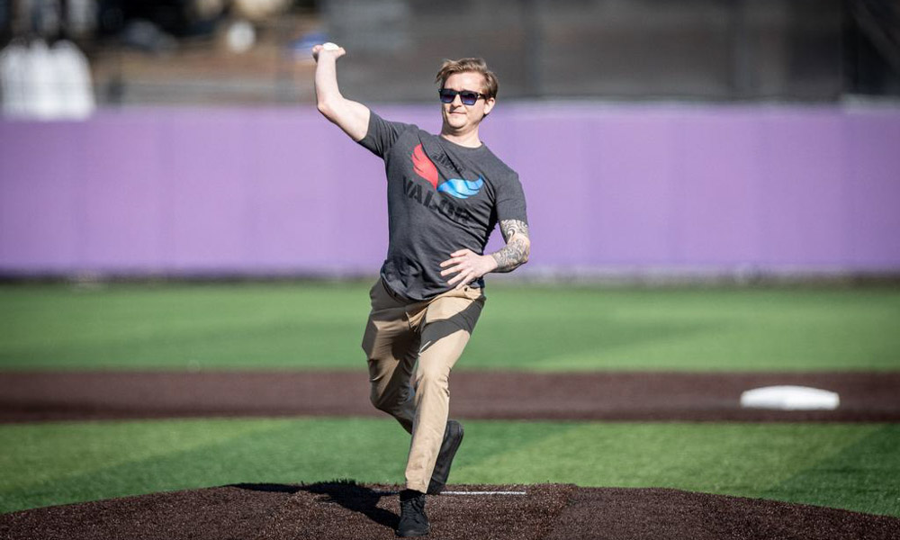 A photo of a male individual wearing a "VALOR" gray t-shirt while throwing out a first pitch at a baseball game.