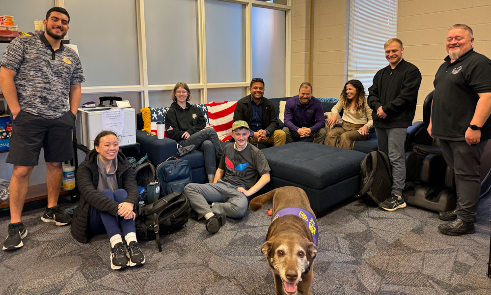 A group photo of individuals and an elderly dog who are all sitting or standing around a couch in the JMU VALOR Resource Center.