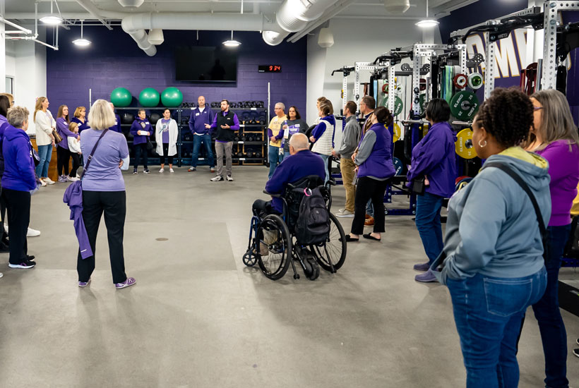 A group of people listening to a speaker in a weight training facility, with fitness equipment visible in the background.