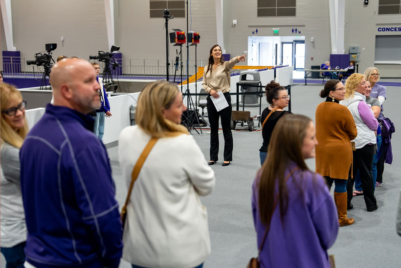 A group of people gathered in a venue, with Lauren Steinbrecher gesturing while speaking, surrounded by media equipment in the background.