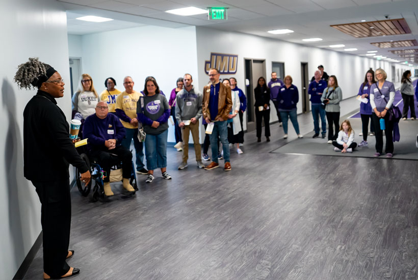 A group of people gathered in a hallway, attentively listening to coach Delethea Quarles, with some participants wearing purple attire representing James Madison University.